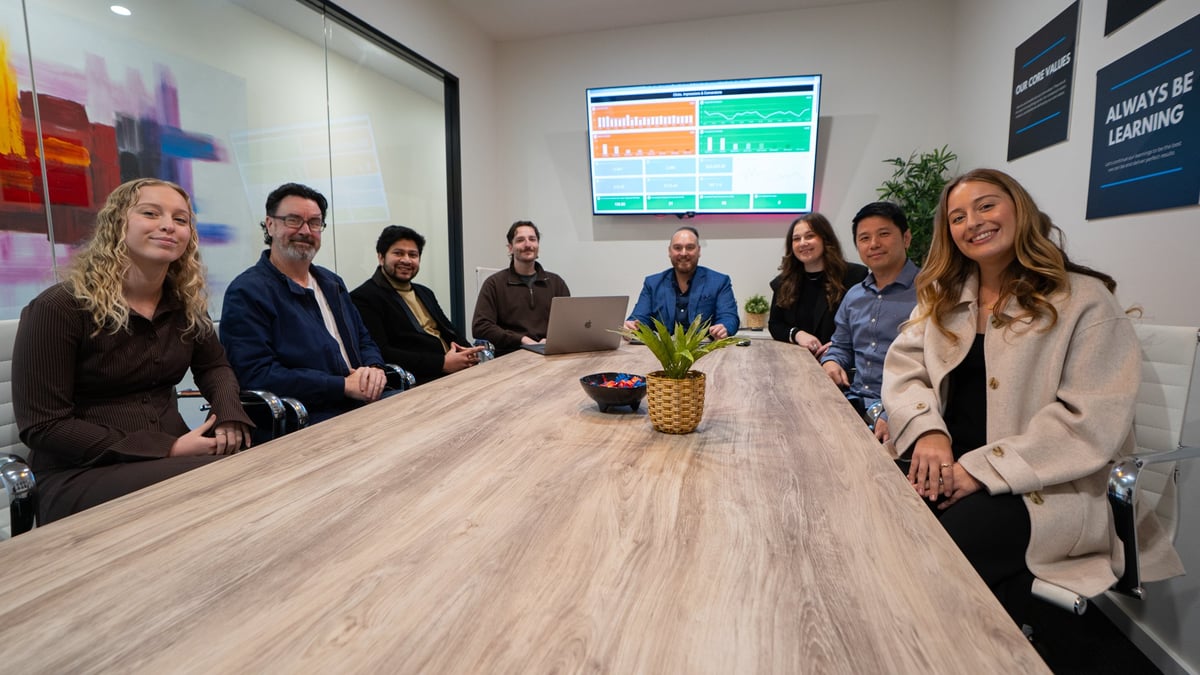 Group of eight business professionals sitting around a wooden conference table in a modern office meeting room with data displays on the wall behind them.