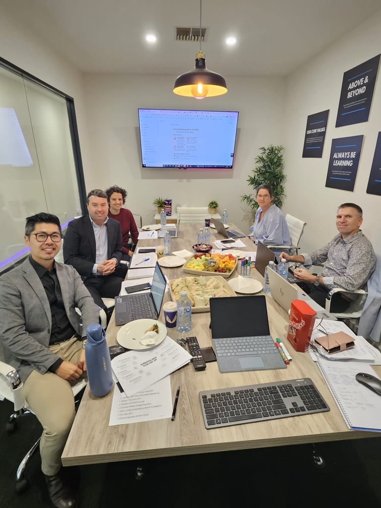 AI training workshops in Adelaide: Five people in business casual attire seated at a conference table with laptops, food, and drinks during a meeting in a modern office