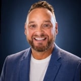 Professional headshot of a smiling man with blonde hair and beard wearing a blue blazer and white shirt against a blue background