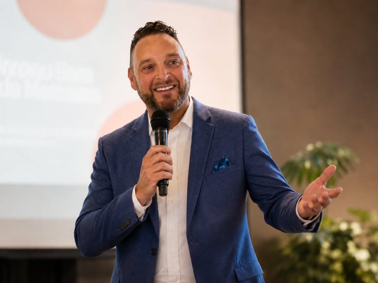Man in blue blazer speaking at microphone during presentation with projection screen visible behind him