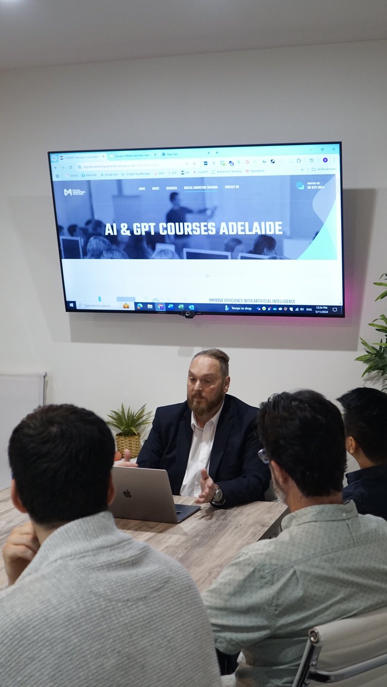 Man in blazer presenting to three people seated at a table with laptop, with AI and GPT Courses Adelaide displayed on wall-mounted screen